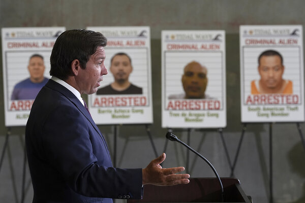 Gov. Ron DeSantis speaks to reporters. Next to him are large posters of suspected criminals caught in recent immigration strikes..